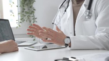 Laboratuvar önlüğü ve steteskop giymiş bir erkek doktor modern tıp ofisinde tablet kullanan hastaya teşhis koymayı açıklarken el kol hareketi yapıyor. İlaç ve sağlık hizmetleri kavramı.
