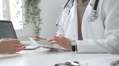 Laboratuvar önlüğü ve steteskop giymiş erkek doktor modern tıp ofisinde tablet kullanırken hastayı dinliyor. İlaç ve sağlık hizmetleri kavramı.