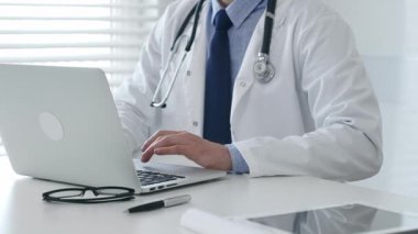 Laboratuvar önlüğü ve steteskop giyen tıp doktoru masasında oturuyor ve güneş gözlüğü ve kalemle yakın bir yerde, parlak bir tıp ofisinde dizüstü bilgisayarda yazıyor. İlaç ve sağlık hizmetleri kavramı