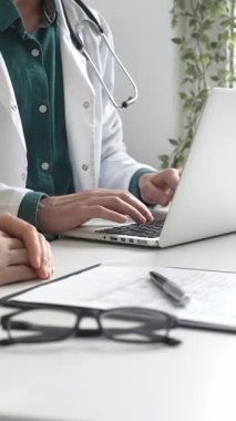 Yeşil gömleğin üzerine laboratuvar önlüğü giyen doktor dizüstü bilgisayarda yazı yazıyor ve klinikteki tıbbi muayene sırasında bir kadının semptomları açıklamasını dinliyor. İlaç ve sağlık hizmetleri kavramı.