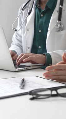 Yeşil gömleğin üzerine laboratuvar önlüğü giyen doktor dizüstü bilgisayarda yazı yazıyor ve klinikteki tıbbi muayene sırasında bir kadının semptomları açıklamasını dinliyor. İlaç ve sağlık hizmetleri kavramı.