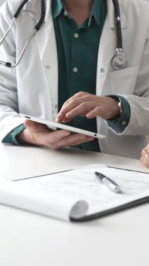 Yeşil gömlekli bir erkek doktor ve modern tıp ofisinde tablet kullanan hastaya teşhisi açıklarken steteskop hareketi yapıyor. İlaç ve sağlık hizmetleri kavramı.