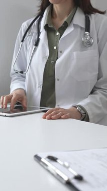 Beyaz laboratuvar önlüğü ve steteskop giyen profesyonel kadın doktor, klinik ortamdaki hasta kayıtlarını incelerken dijital tablet üzerinde çalışıyor. İlaç ve sağlık hizmetleri kavramı