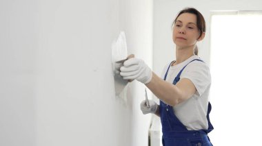 Profesyonel inşaat işçisi kadın, beyaz bir duvarı onarmak için macun bıçağı kullanıyor, bir kat macun sürüyor ve temiz bir iş için beyaz eldiven takıyor.