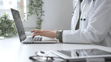 Steteskop ve laboratuvar önlüğü giyen doktor kadın özenle bir dizüstü bilgisayarda yazıyor ve modern bir tıp ofisinde etkin bir sağlık hizmeti yönetimi sergiliyor. İlaç ve sağlık hizmetleri.