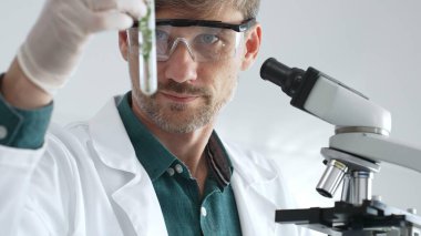 Maskeli yetişkin bir bilim adamı ve yeşil bitki örneğiyle test tüpü tutan eldivenler mikroskop ile laboratuvar araştırması yapıyor. Tıp, sağlık ve bilim konsepti.