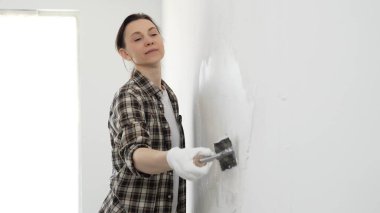 Koruyucu eldiven ve kareli gömlek giyen kadın, iç dekorasyon çalışmalarını boyamadan önce hasarlı bölgeleri yumuşatmak için beyaz duvar yüzeyine macun sürüyor..