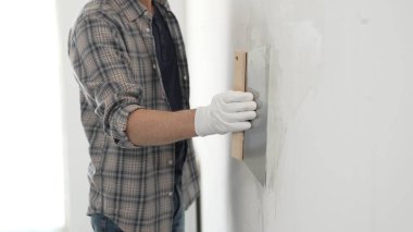 Bilinmeyen erkek inşaat işçisi koruyucu eldivenler ve ekose gömlek giyiyor. Tadilat projesi sırasında profesyonel alçıpan bantlama bıçağını kullanarak duvarın yüzeyine sorunsuzca alçı yayıyor..