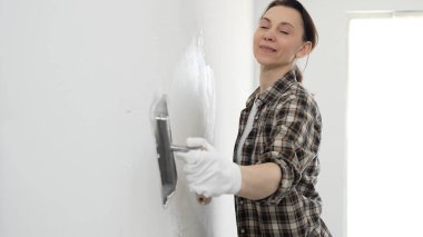 Koruyucu eldiven ve kareli gömlek giyen kadın, iç dekorasyon çalışmalarını boyamadan önce hasarlı bölgeleri yumuşatmak için beyaz duvar yüzeyine macun sürüyor..