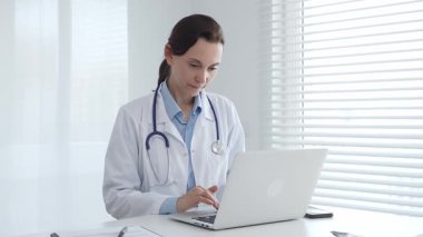 Laboratuvar önlüğü ve steteskop giyen kendine güvenen kadın doktor, doğal ışık dolu, parlak ve modern bir muayenehanede çalışırken laptopuna odaklanmış bir şekilde bir masada oturuyor.