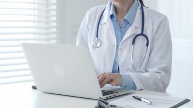 Laboratuvar önlüğü ve steteskop giyen kadın doktor masamda oturmuş bilgisayarında tıbbi belgeler ve kalemle ön planda yazı yazıyor, arka planda panjurları olan parlak bir pencere var.