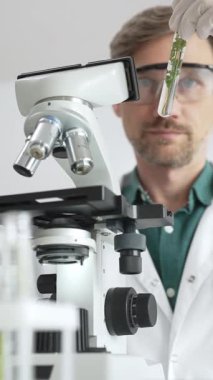 Koruyucu giysiler giyen bilim adamı mikroskop, cam ve bilimsel ekipman yakınında yeşil bitki örnek tüpünü analiz ediyor. Tıp ve bilim konsepti.