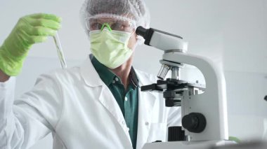 Yeşil koruyucu ekipman giyen yetişkin bir bilim adamı test tüplerindeki bitki örneklerini analiz ediyor ve botanik araştırmaları için modern bir laboratuvarda mikroskop kullanıyor. Tıp, sağlık ve bilim konsepti.