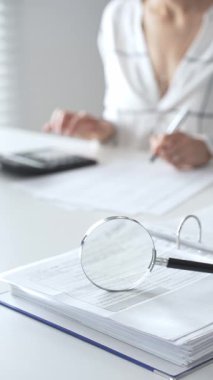 Finansal belgelerde büyüteç odak noktasındayken, hesap makinesi kullanan ve notlar alan bir muhasebeci dikey görüşü kapatıyor. Ticarette denetim ve vergiler.