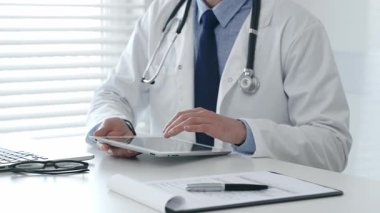 Dijital tablet üzerinde çalışan, beyaz laboratuvar önlüğü ve steteskop giyen, dizüstü bilgisayarı, gözlüğü ve hasta tablosu olan bir tıbbi uzman. İlaç ve sağlık hizmetleri kavramı