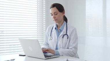 Laboratuvar önlüğü ve steteskop giyen kendine güvenen kadın doktor, doğal ışık dolu, parlak ve modern bir muayenehanede çalışırken laptopuna odaklanmış bir şekilde bir masada oturuyor.