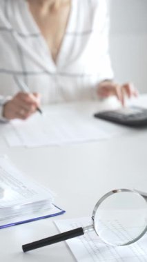 Finansal belgelerde büyüteç odak noktasındayken, hesap makinesi kullanan ve notlar alan bir muhasebeci dikey görüşü kapatıyor. Ticarette denetim ve vergiler.