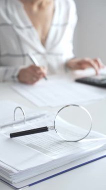 Finansal belgelerde büyüteç odak noktasındayken, hesap makinesi kullanan ve notlar alan bir muhasebeci dikey görüşü kapatıyor. Ticarette denetim ve vergiler.