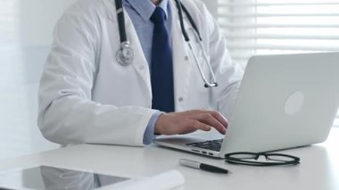 Laboratuvar önlüğü ve steteskop giyen tıp doktoru masasında oturuyor ve güneş gözlüğü ve kalemle yakın bir yerde, parlak bir tıp ofisinde dizüstü bilgisayarda yazıyor. İlaç ve sağlık hizmetleri kavramı