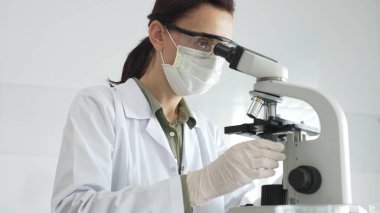 Modern laboratuvar ortamında biyolojik örnekleri incelerken mikroskoba odaklanan koruyucu ekipman takan kadın bilim adamı. Tıp, sağlık ve bilim konsepti