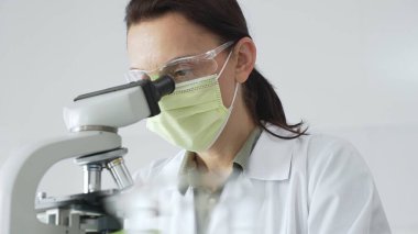 Örnek inceleyen profesyonel kadın bilim adamı laboratuvar çalışma alanında yeşil koruyucu giysi giyerken mikroskop altına kayıyor. Tıp, sağlık ve bilim konsepti.