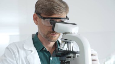 Koruyucu gözlük ve laboratuvar önlüğü takan yetişkin bir bilim adamı modern bir laboratuvar ortamında mikroskopla örnekleri analiz ediyor. Tıp, sağlık ve bilim konsepti.