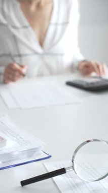 Finansal belgelerde büyüteç odak noktasındayken, hesap makinesi kullanan ve notlar alan bir muhasebeci dikey görüşü kapatıyor. Ticarette denetim ve vergiler.