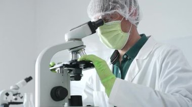 Koruyucu maske, kep ve yeşil eldiven takan tıp bilimcileri mikroskop kullanarak laboratuvarda bilimsel araştırmalar yapıyor, aşı veya ilaç geliştiriyor. Tıp, sağlık ve bilim konsepti.