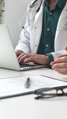 Yeşil gömleğin üzerine laboratuvar önlüğü giyen doktor dizüstü bilgisayarda yazı yazıyor ve klinikteki tıbbi muayene sırasında bir kadının semptomları açıklamasını dinliyor. İlaç ve sağlık hizmetleri kavramı.