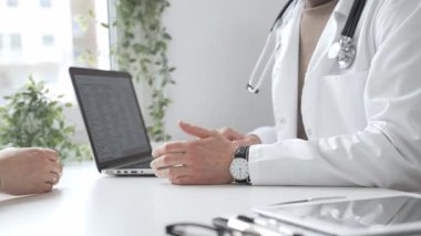 Laboratuvar önlüklü ve steteskoplu doktor, tıbbi ofisindeki dizüstü bilgisayarla hastaya teşhis koymayı açıklarken el kol hareketi yapıyor. İlaç ve sağlık hizmetleri kavramı.