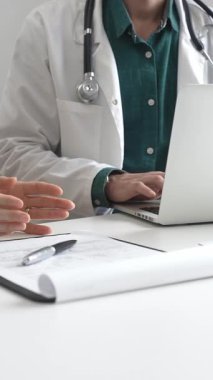 Yeşil gömleğin üzerine laboratuvar önlüğü giyen doktor dizüstü bilgisayarda yazı yazıyor ve klinikteki tıbbi muayene sırasında bir kadının semptomları açıklamasını dinliyor. İlaç ve sağlık hizmetleri kavramı.