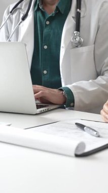 Yeşil gömleğin üzerine laboratuvar önlüğü giyen doktor dizüstü bilgisayarda yazı yazıyor ve klinikteki tıbbi muayene sırasında bir kadının semptomları açıklamasını dinliyor. İlaç ve sağlık hizmetleri kavramı.