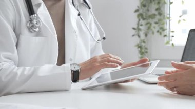 Laboratuvar önlüğü ve steteskop giymiş erkek doktor modern tıp ofisinde tablet kullanırken hastayı dinliyor. İlaç ve sağlık hizmetleri kavramı.