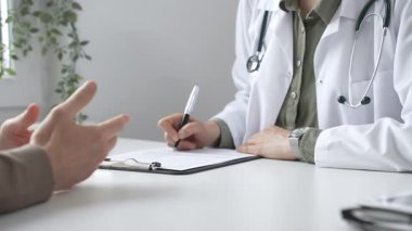 Doktor kadın, erkek hasta sağlığıyla ilgili endişeleri dinliyor ve parlak, modern bir klinikteki muayenede reçete yazıyor. Kapatın. İlaç ve sağlık hizmetleri kavramı.