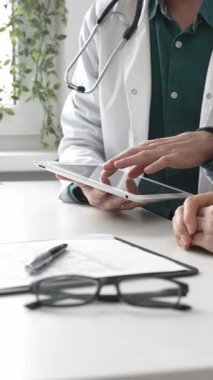 Yeşil gömlekli bir erkek doktor ve modern tıp ofisinde tablet kullanan hastaya teşhisi açıklarken steteskop hareketi yapıyor. İlaç ve sağlık hizmetleri kavramı.