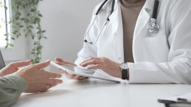 Laboratuvar önlüğü ve steteskop giymiş erkek doktor modern tıp ofisinde tablet kullanırken hastayı dinliyor. İlaç ve sağlık hizmetleri kavramı.