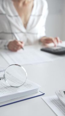 Finansal belgelerde büyüteç odak noktasındayken, hesap makinesi kullanan ve notlar alan bir muhasebeci dikey görüşü kapatıyor. Ticarette denetim ve vergiler.