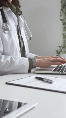 Stetoskop ve laboratuvar önlüğü giyen doktor özenle bir dizüstü bilgisayarda yazıyor ve modern bir tıp ofisinde verimli bir sağlık hizmeti yönetimi sergiliyor. İlaç ve sağlık hizmetleri kavramı