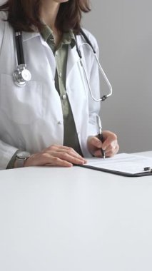 Laboratuvar önlüğü ve steteskop giymiş kadın doktor, tıp bürosunun masasındaki panoya profesyonelliği vurgulayan dizüstü bilgisayar ve gözlüklerle notlar yazıyor. İlaç ve sağlık hizmetleri kavramı