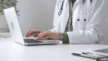 Stetoskop ve laboratuvar önlüğü giyen doktor özenle bir dizüstü bilgisayarda yazıyor ve modern bir tıp ofisinde verimli bir sağlık hizmeti yönetimi sergiliyor. İlaç ve sağlık hizmetleri kavramı