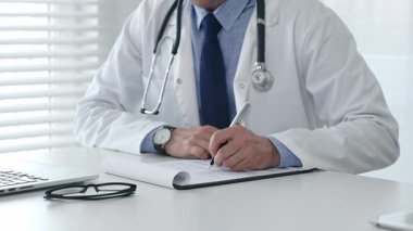 Laboratuvar önlüğü ve steteskop giyen erkek doktor, tıbbi bir ofiste bir masada oturuyor, hasta bakımı ve tedavisi için bir not panosuna not alırken dizüstü bilgisayar kullanmaya odaklanıyor.