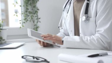 Tablet üzerindeki hasta verilerini inceleyen, çağdaş klinik ortamlarda beyaz laboratuvar önlüğü ve steteskop giyen, dijital sağlık danışmanlığı yapan bir tıp uzmanı. İlaç ve sağlık hizmetleri