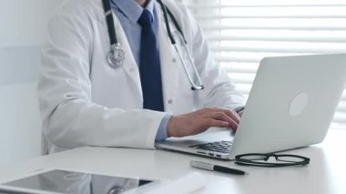 Laboratuvar önlüğü ve steteskop giyen tıp doktoru masasında oturuyor ve güneş gözlüğü ve kalemle yakın bir yerde, parlak bir tıp ofisinde dizüstü bilgisayarda yazıyor. İlaç ve sağlık hizmetleri kavramı