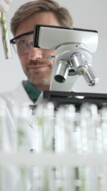 Koruyucu giysiler giyen bilim adamı mikroskop, cam ve bilimsel ekipman yakınında yeşil bitki örnek tüpünü analiz ediyor. Tıp ve bilim konsepti.