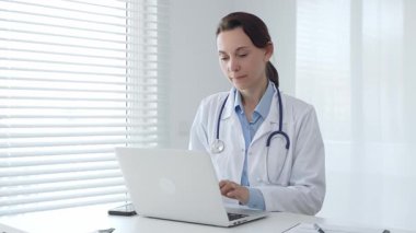 Laboratuvar önlüğü ve steteskop giyen kendine güvenen kadın doktor, doğal ışık dolu, parlak ve modern bir muayenehanede çalışırken laptopuna odaklanmış bir şekilde bir masada oturuyor.