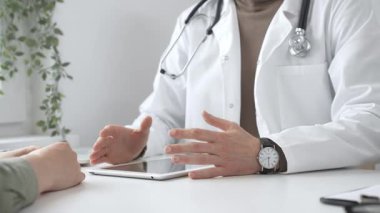Laboratuvar önlüğü ve steteskop giymiş bir erkek doktor modern tıp ofisinde tablet kullanan hastaya teşhis koymayı açıklarken el kol hareketi yapıyor. İlaç ve sağlık hizmetleri kavramı.