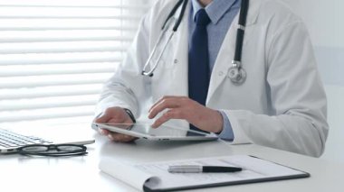 Dijital tablet üzerinde çalışan, beyaz laboratuvar önlüğü ve steteskop giyen, dizüstü bilgisayarı, gözlüğü ve hasta tablosu olan bir tıbbi uzman. İlaç ve sağlık hizmetleri kavramı