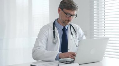 Gözlük takan ve steteskoplu laboratuvar önlüğü giyen olgun bir erkek doktor, parlak sağlık ofisindeki masada otururken dizüstü bilgisayarında klavye kullanıyor.