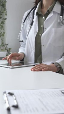 Beyaz laboratuvar önlüğü ve steteskop giyen profesyonel kadın doktor, klinik ortamdaki hasta kayıtlarını incelerken dijital tablet üzerinde çalışıyor. İlaç ve sağlık hizmetleri kavramı