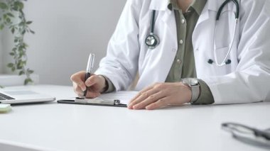 Laboratuvar önlüğü ve steteskop giymiş kadın doktor, tıp bürosunun masasındaki panoya profesyonelliği vurgulayan dizüstü bilgisayar ve gözlüklerle notlar yazıyor. İlaç ve sağlık hizmetleri kavramı